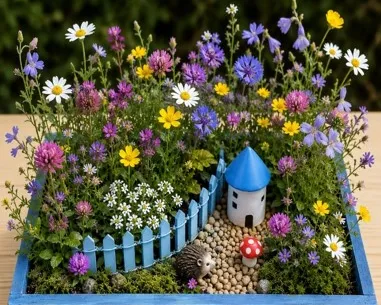 Miniaturgarten in einer blauen Kiste mit bunten Blumen, kleinem Haus, Zaun, Weg und Dekorfiguren wie Igel und Pilz.