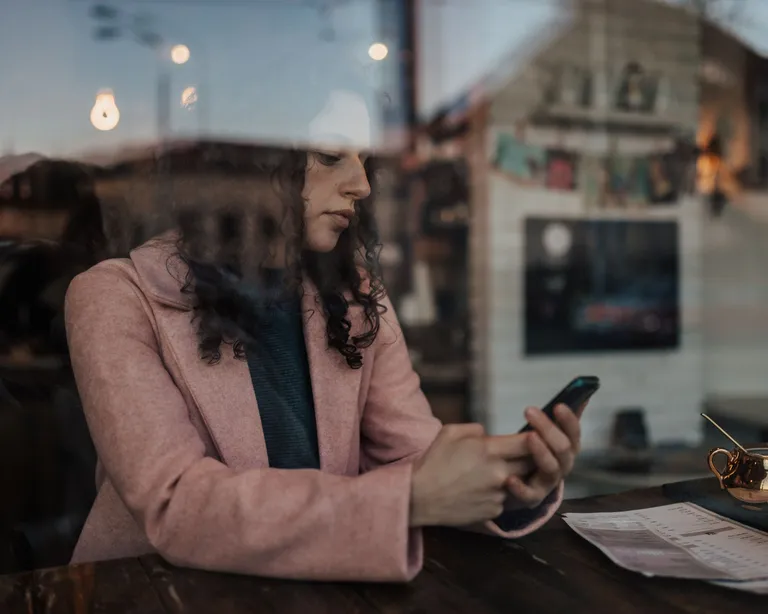Frau sitzt in einem Café am Fenster, trägt einen rosa Mantel und schaut auf ihr Smartphone, draußen spiegelt sich die Stadt.