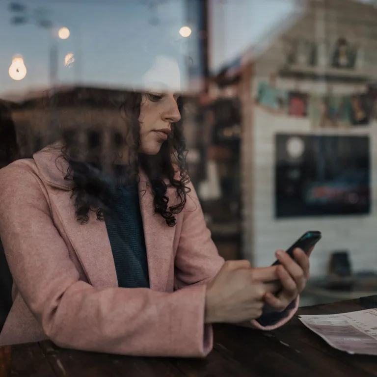 Frau sitzt in einem Café am Fenster, trägt einen rosa Mantel und schaut auf ihr Smartphone, draußen spiegelt sich die Stadt.