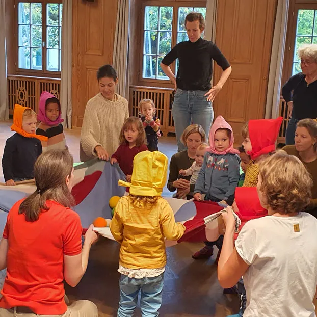 Mehrere Kinder und Erwachsene spielen gemeinsam in einem Raum, einige Kinder tragen bunte Hüte, alle sitzen oder stehen im Kreis.