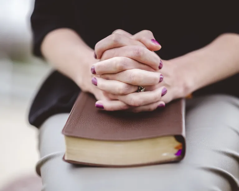 Verschränkte Hände liegen auf einem geschlossenen Buch, vermutlich einer Bibel, im Schoss einer sitzenden Person.