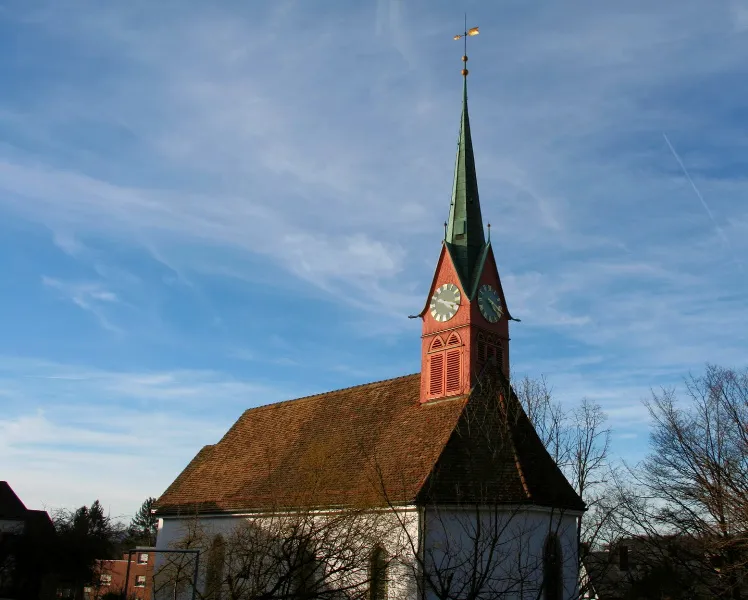 Kirche mit rotem Turm und grünem spitzen Dach unter blauem Himmel, umgeben von Bäumen.