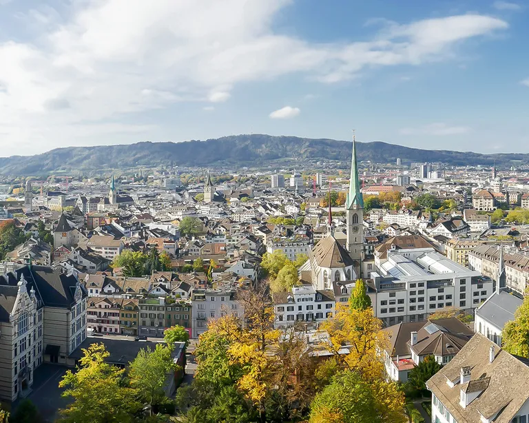 Panorama der Stadt Zürich mit vielen Häusern, einer Kirche mit hohem Turm und umliegenden Hügeln unter blauem Himmel.